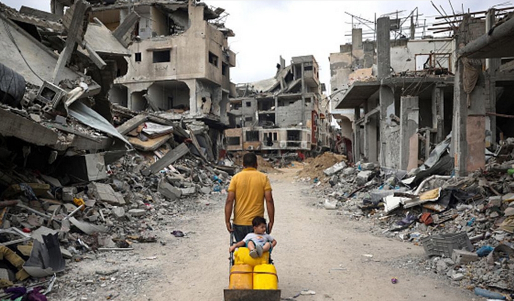 فلسطيني يجر طفله الجالس فوق غالونات المياه وسط الدمار في غزة